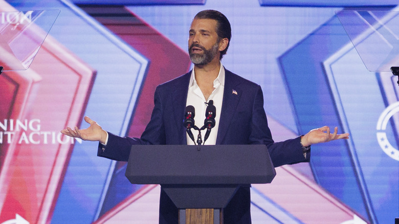 Donald Trump Jr. speaking onstage at AmericaFest 2025 in a navy blazer and open-collared white shirt