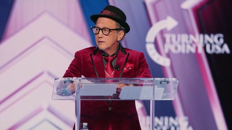 Rob Schneider onstage at AmericaFest 2025 in a garish red suit