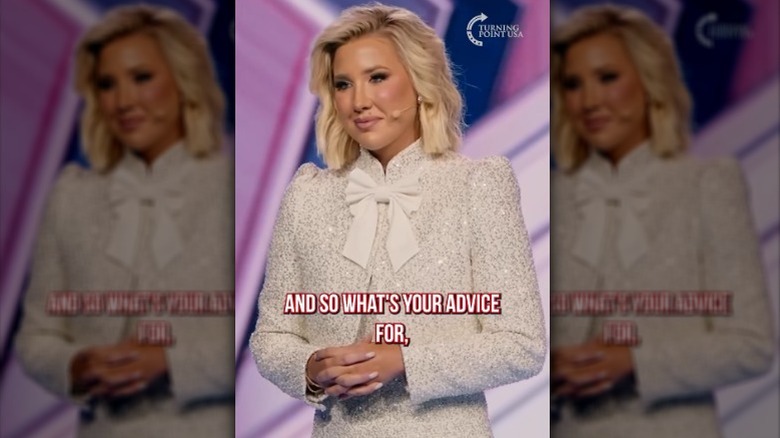 Savannah Chrisley speaking onstage in a white suit at AmericaFest 2025