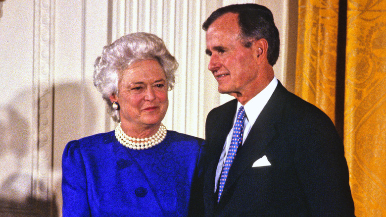 Barbara Bush in a blue dress with pearls standing next to George H .W. Bush in a black suit