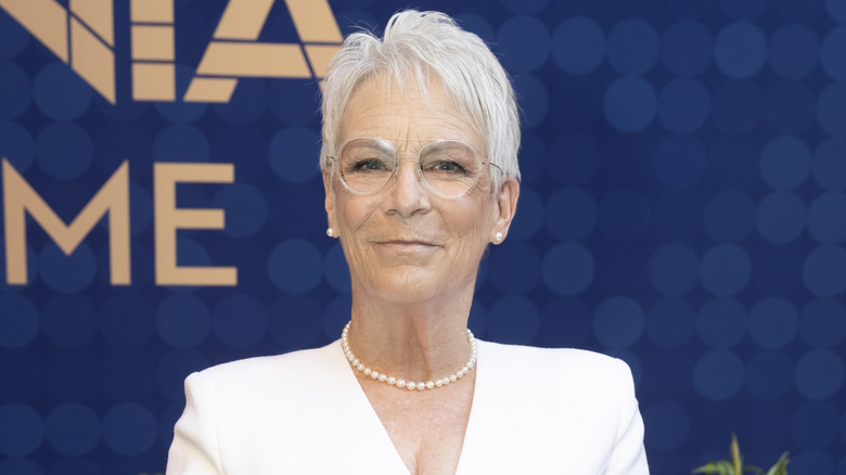 Jamie Lee Curtis short gray bob, glasses, pearls and a white dress in front of a dark blue background