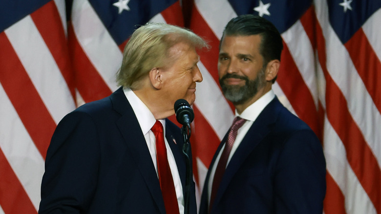 Donald Trump Jr. and Donald Trump smiling behind a podium in front of an American flag