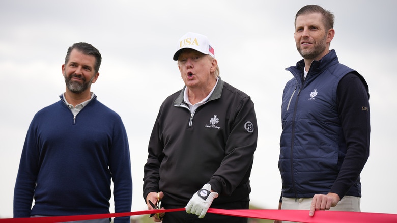 Donald Trump about to cut a red ribbon flanked by Don Jr. and Eric Trump