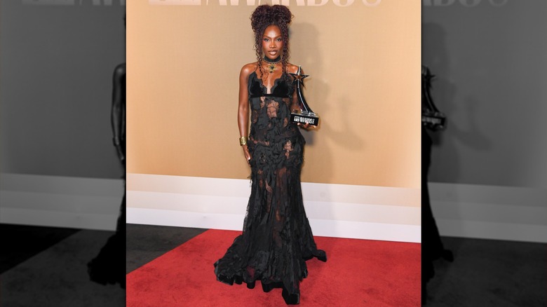 Doechii, in a black dress, holding her BET Award on the red carpet in 2026