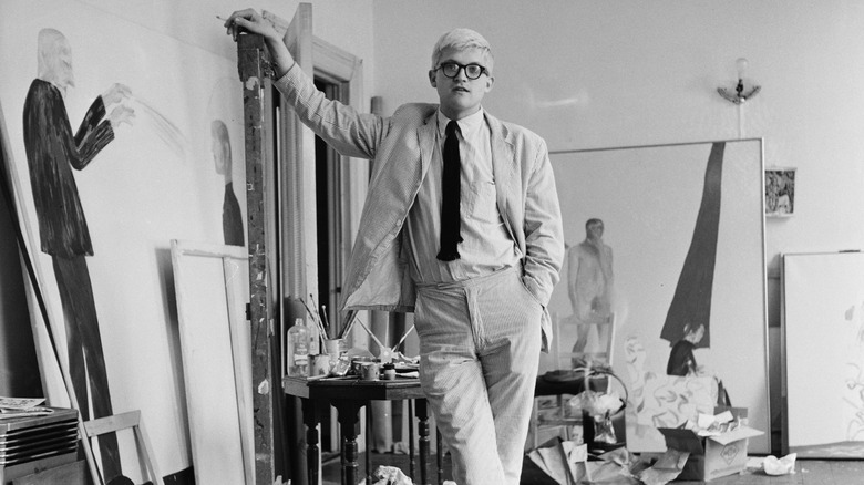David Hockney in his studio, surrounded by paintings, wearing a pinstripe suit.
