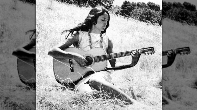 Joan Baez sitting in a field and playing her guitar in 1965.