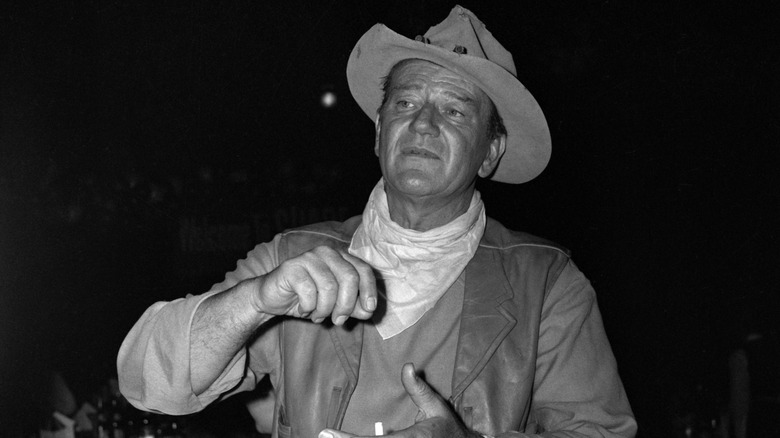 John Wayne in a hat, neckerchief and leather vest at a dinner event in the 1960s.