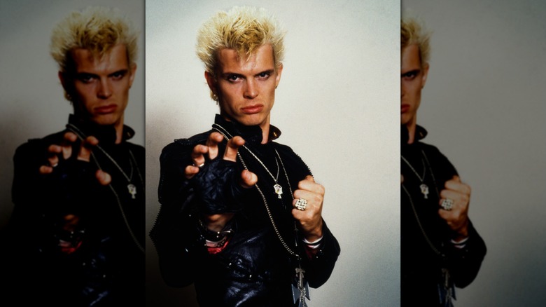Billy Idol sneering in 1984, wearing all black with heavy accessories.