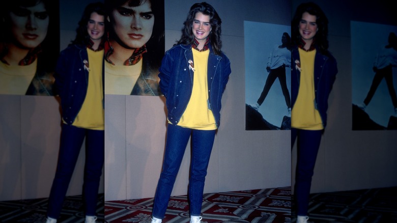 Brooke Shields smiling in 1985 while wearing a jean jacket and pants set with a yellow shirt.