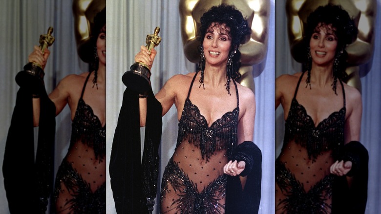 Cher holding up her Oscar at the 60th Academy Awards, wearing a black sequined Bob Mackie gown.