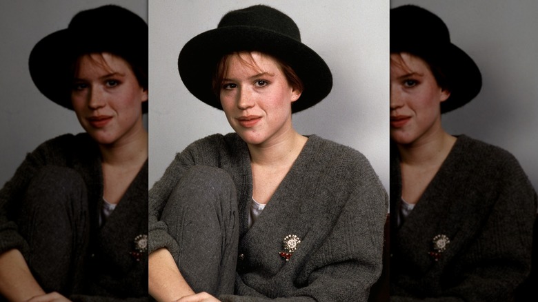 Molly Ringwald half-smiling in 1985, wearing a gray sweater and black hat.