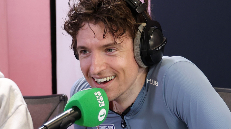 Greg James talking into a radio show microphone while wearing headphones.