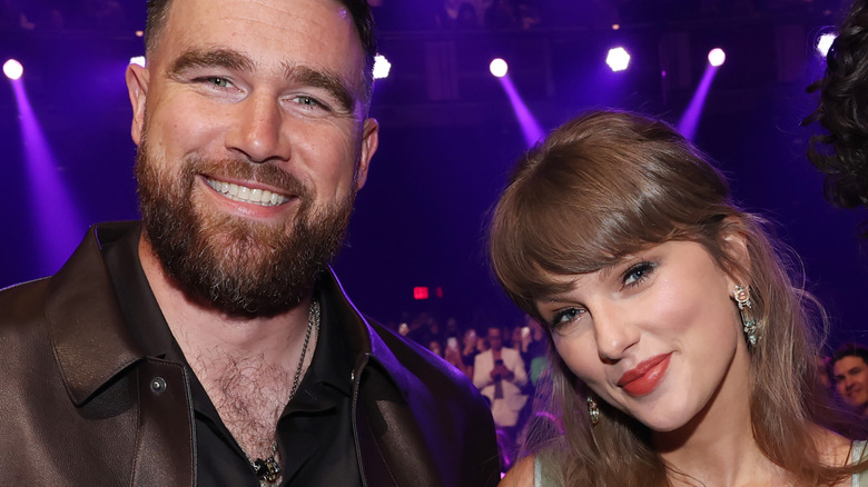 Travis Kelce and Taylor Swift smiling against a purple backdrop.