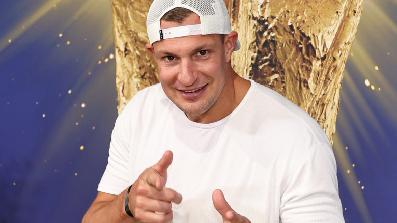 Rob Gronkowski smiling and pointing to camera against a blue and gold backdrop.