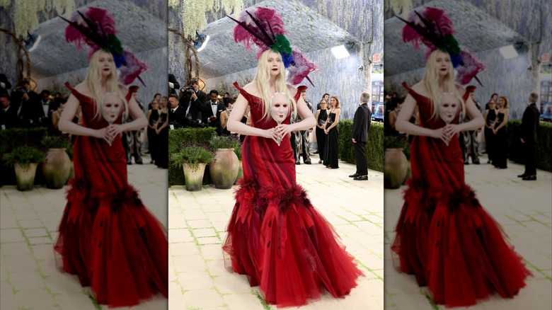 Gwendoline Christie in a large feathered hat, burgundy floor-length gown, and a mask of her own face in-hand