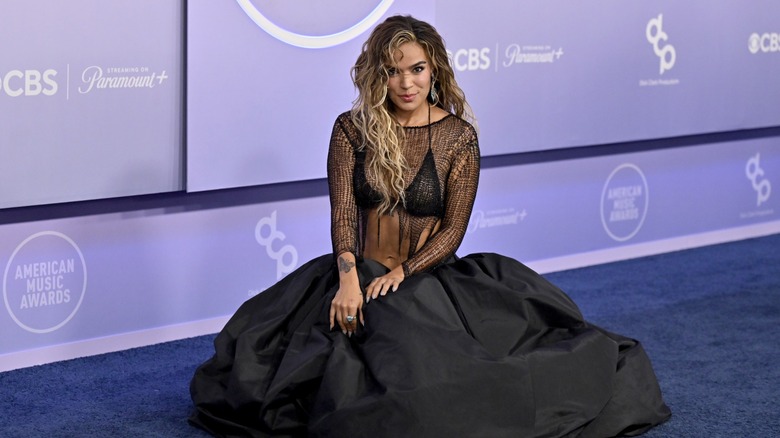Karol G crouched down and smiling on the red carpet of the 2026 AMAs