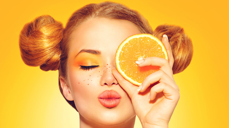 Woman with orange lipstick holding orange slice to her eye