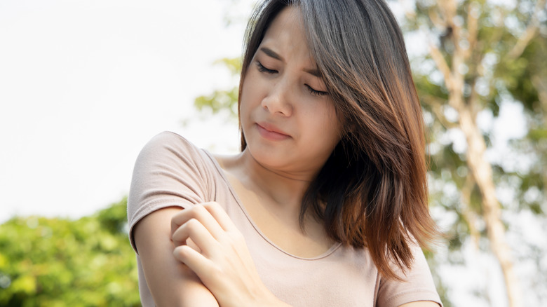 Brunette woman outside scratching her arm.