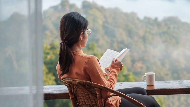 woman sitting in chair & holding book while looking out at scenery. Coffee sits on railing