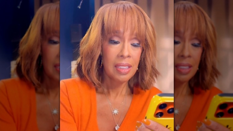 Gayle King wearing fake eyelashes and dark eyeliner and looking at her phone