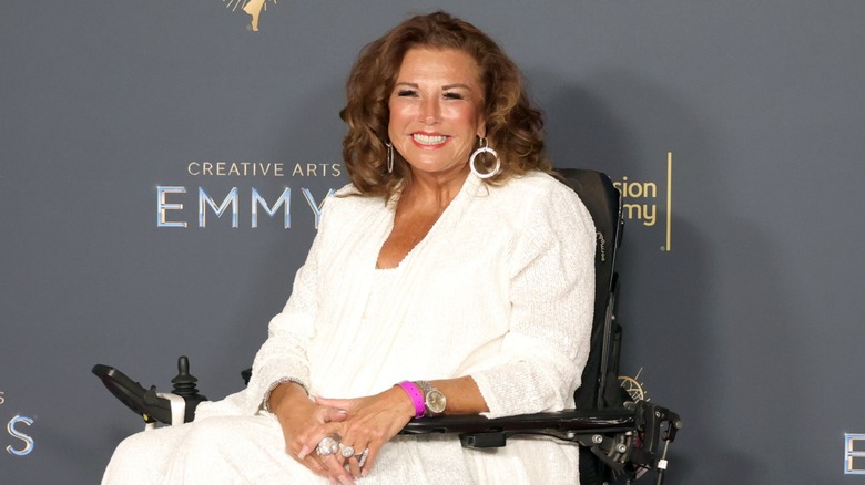 Abby Lee Miller in white pantsuit and hoop earrings in a wheelchair