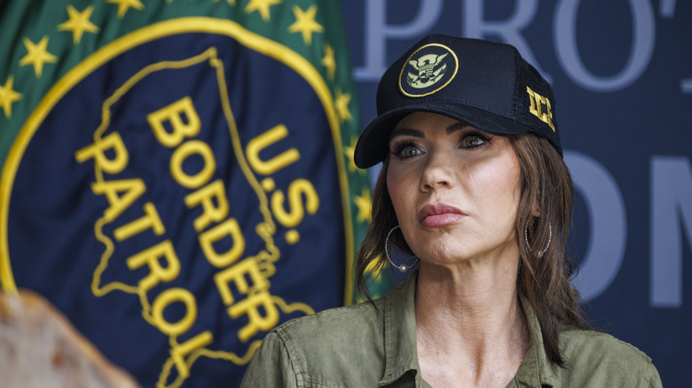 Kristi Noem wearing an ICE hat in front of a U.S. Border Patrol flag