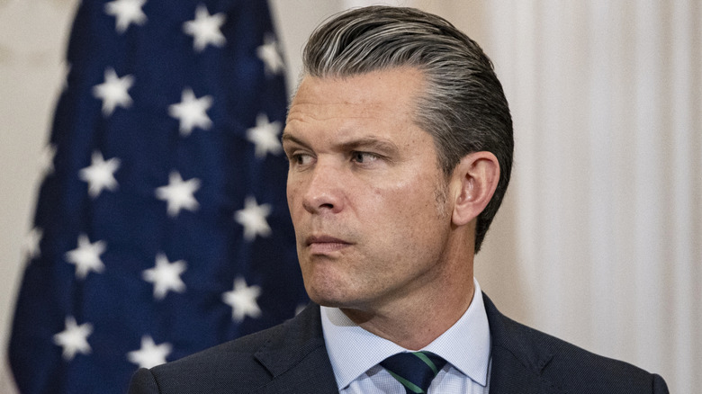 Pete Hegseth raising his eyebrow and pursing his lip in front of an American flag