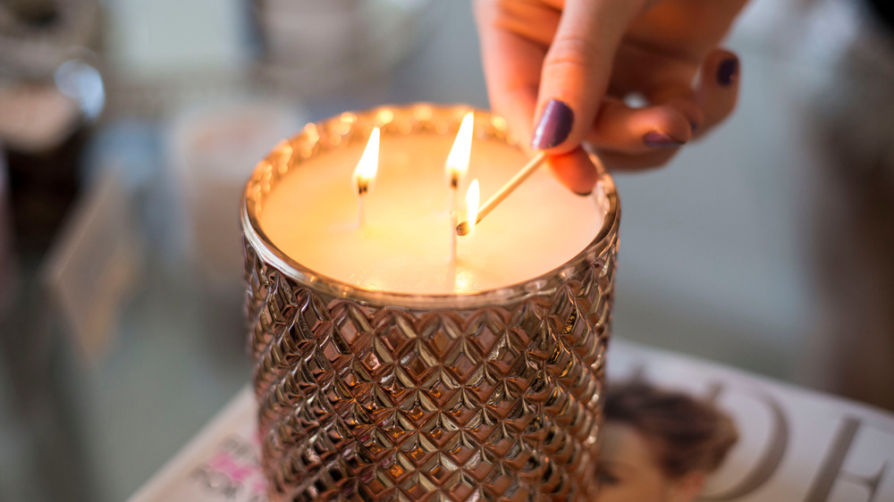 A hand with dark painted fingernails lights a three wick candle in a metallic holder