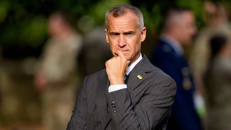 Corey Lewandowski holding a finger to his chin in a grey suit