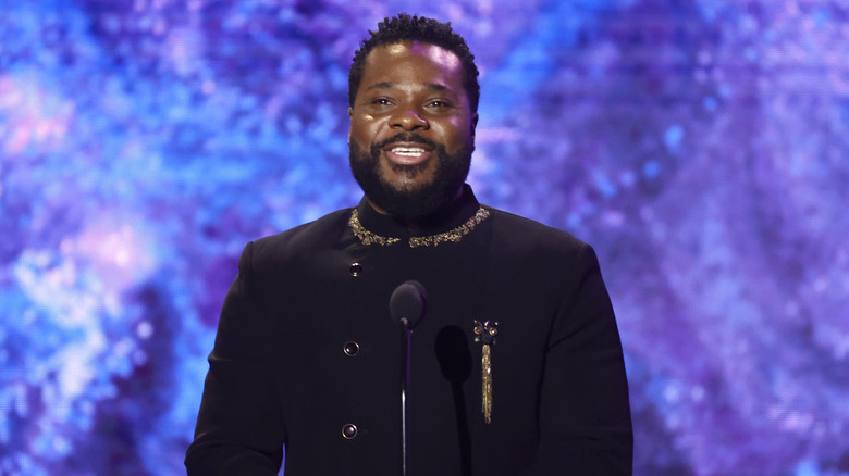 Malcolm-Jamal Warner in black coat with gold trim in front of mic