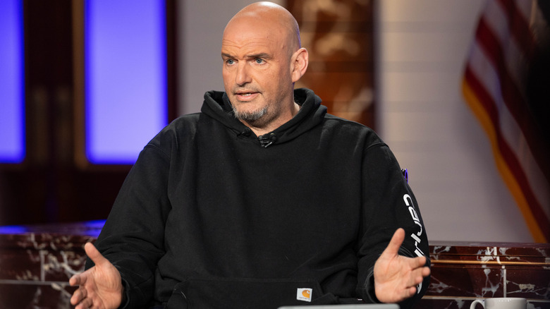John Fetterman speaking onstage in a black hoodie while raising his hands