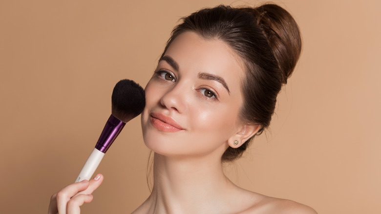 Young woman with her brunette hair in a bun holding a large makeup brush on her cheek.