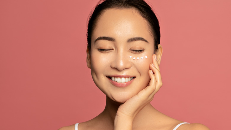 young woman smiling with eyes closed, applying dots of skincare cream under eyes