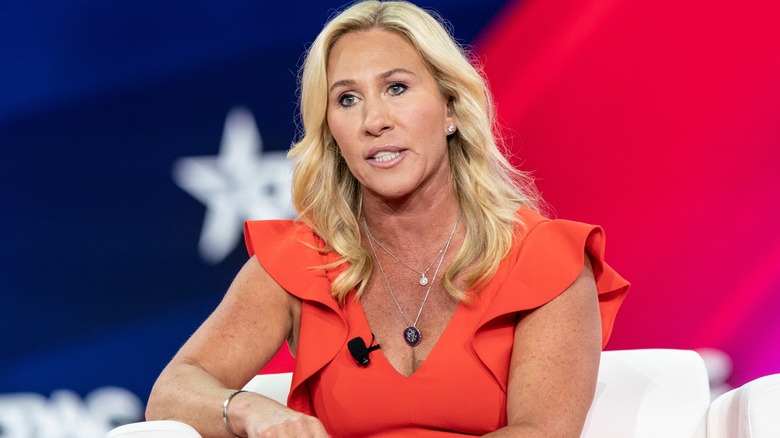 Congresswoman Marjorie Taylor Greene speaks in orange ruffled top with curled hair and silver necklaces and bracelet