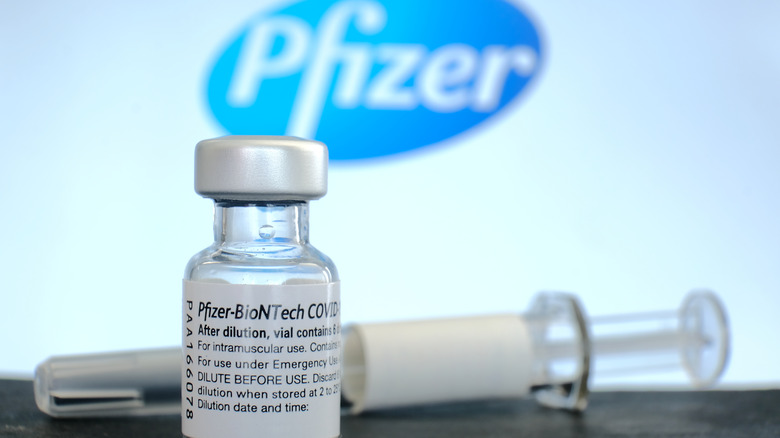 Pfizer covid vaccine on a table with a needle with a Pfizer logo behind it.