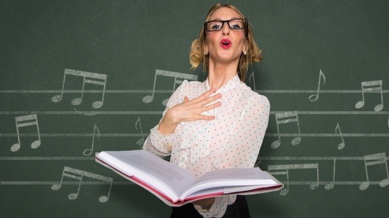 Woman in glasses holding a huge open book and singing in front of musical notes on a chalkboard