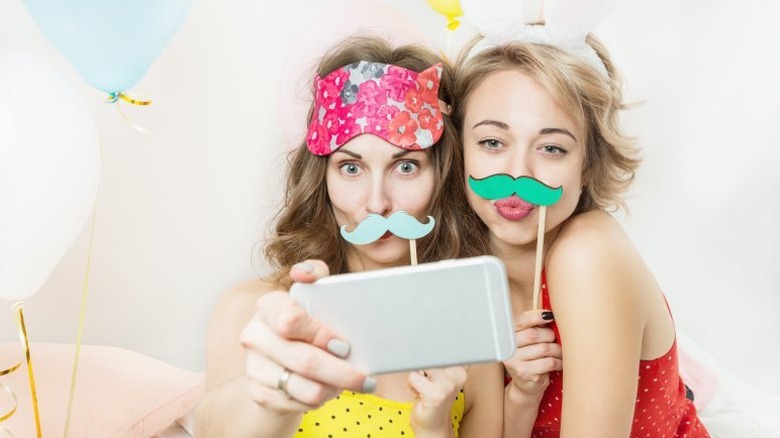 Two young women taking a selfie while holding colorful fake mustaches up to their faces