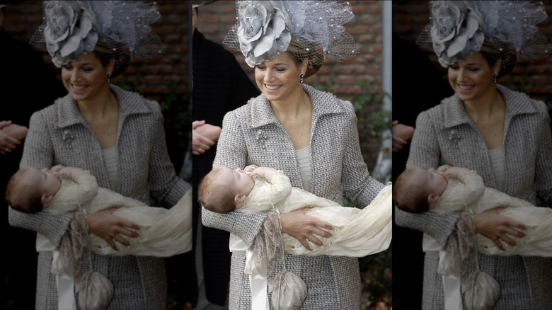 Queen Máxima looks down holding her baby, Princess Alexia, wearing a gray and lilac netted hat with pearls