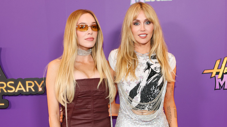 Alex Cooper wears sunglasses and a brown top and poses beside Miley Cyrus, who wears a silver dress and deconstructed "Hannah Montana" shirt.