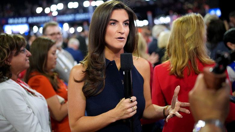 Kaitlan Collins speaking to the camera with a mic in a navy sleeveless dress