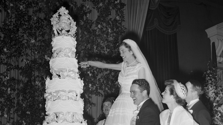 Eunice Kennedy cutting her wedding cake while standing on a ladder in a wedding dress