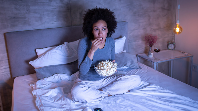 Mixed race woman lit up by off-camera TV screen eating popcorn in bed
