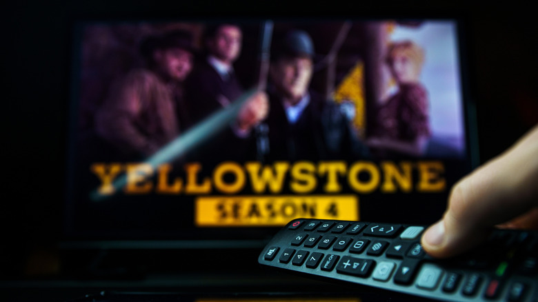 A thumb pressing on a remote control in front of a TV displaying an out-of-focus 'Yellowstone' image