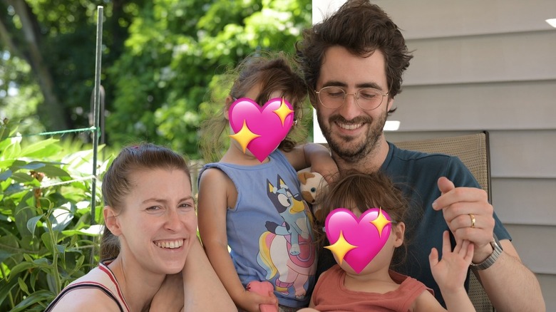 Eden Sher smiles outside next to her husband who holds their twin daughters whose faces are hidden by hearts