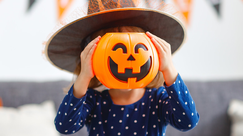 A child hiding their face behind a smiling Halloween candy bucket