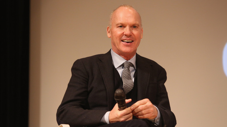 Smiling Michael Keaton wearing formalwear with a mic in his hand.