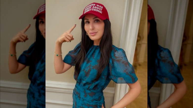 Laura Loomer poses while wearing a blue dress and pointing to a red "MAGA" style hat that reads "Loomer was right about everything"