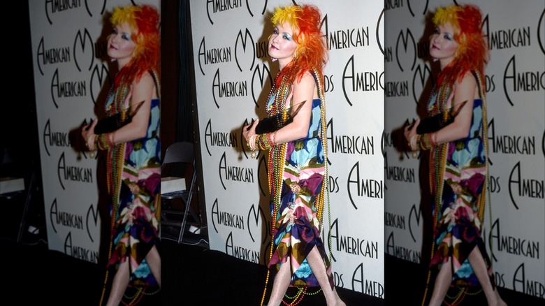 Cyndi Lauper with yellow and red hair wearing colorful gown with beads in standing in front of AMA wall
