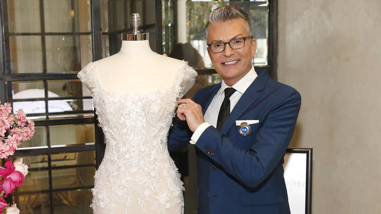 Randy Fenoli smiling next to a wedding dress on a mannequin
