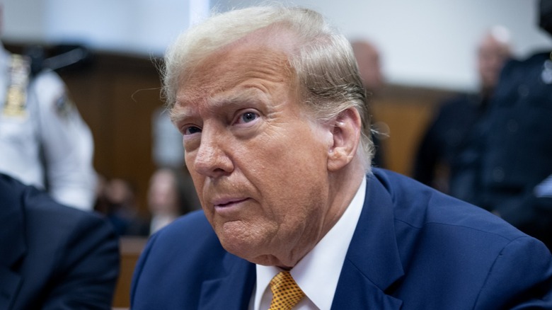 Donald Trump sitting in court on trial for his conduct with Stormy Daniels in a blue suit and yellow tie